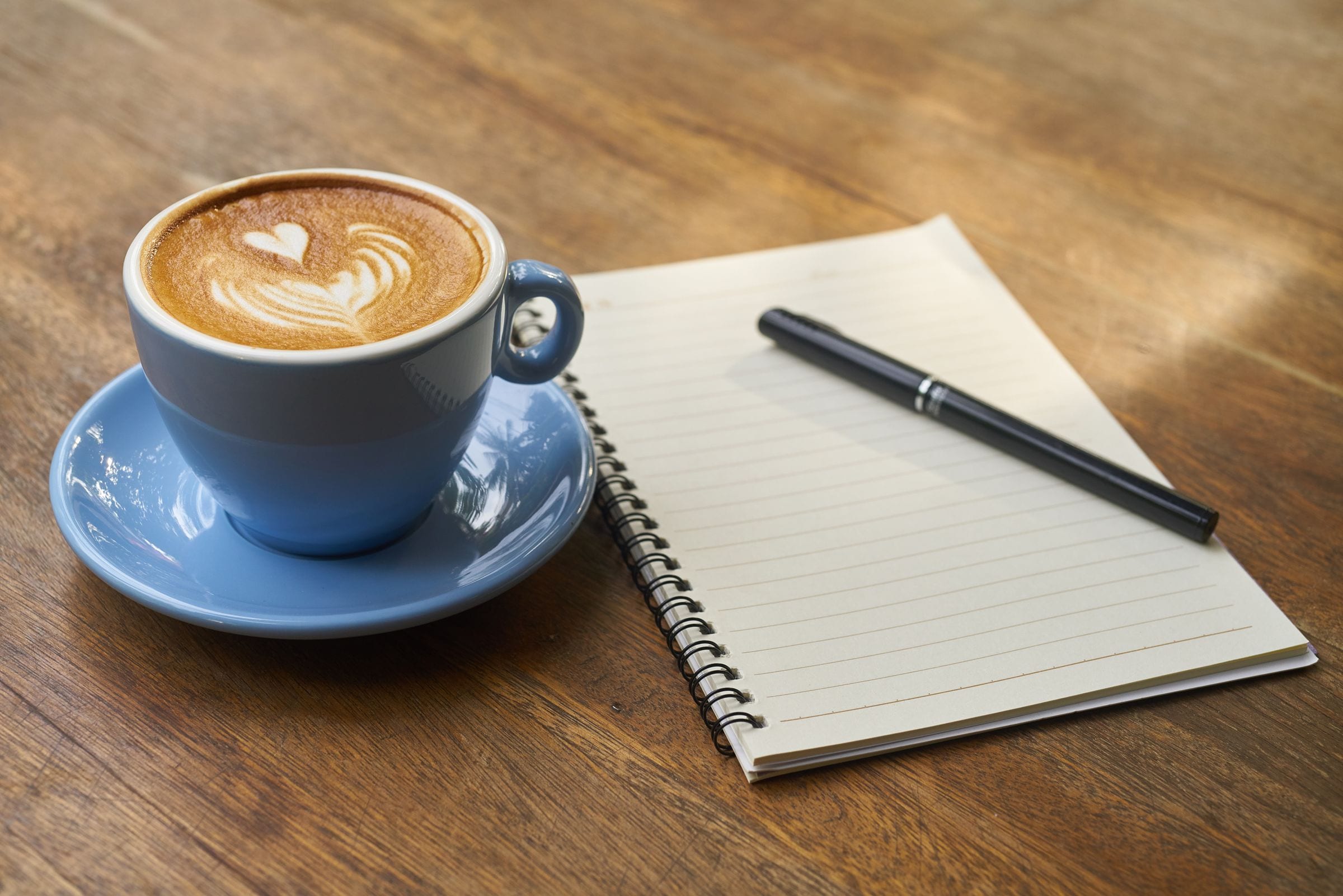 An open notebook with a pen and a cup of latte on a wooden table